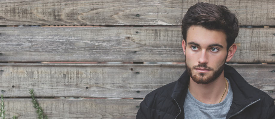 young brunette man with mustache and beard leaning against wood wall