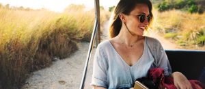 brunette woman wearing sunglasses sitting in cart, traveling, beach and grass behind her