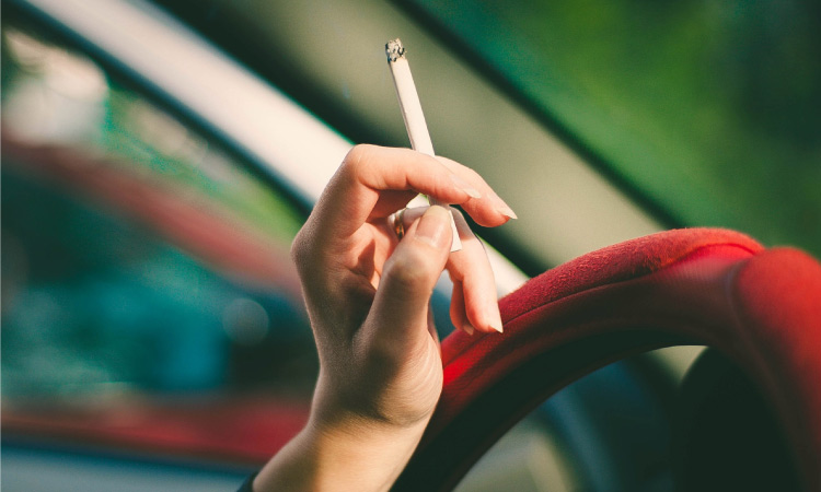 A manicured hand holding a cigarette rests on a steering wheel with a red cover