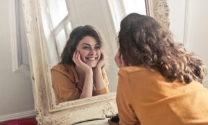 Brunette young woman wearing yellow blouse looks in the mirror and smiles excitedly after her dental appointment