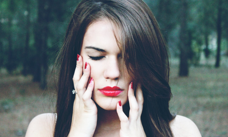 Closeup of brunette woman with red lipstick and fingernails touching her cheeks due to tooth sensitivity and pain